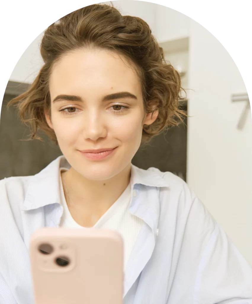 A young woman with short, curly hair smiles gently while looking at her smartphone. She is dressed casually in a light blue shirt over a white top. The background is a modern, minimalistic setting, creating a relaxed atmosphere as she engages with her device.