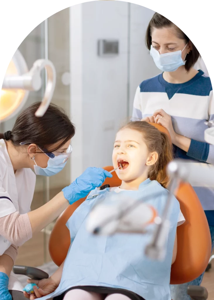 A young girl sits in a dental chair, smiling with her mouth open as a dental professional examines her teeth with a tool. The professional wears a mask and protective eyewear. In the background, a parent stands nearby, also wearing a mask, observing the appointment in a modern dental office.