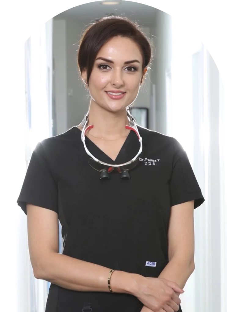 Dr. Parisa, a smiling dental professional, stands confidently in a modern dental office wearing a black scrubs top with her name and credentials displayed. She has a dental loupe around her neck and is posed with her arms crossed, exuding warmth and professionalism.