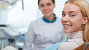 A smiling young woman in a dental chair, wearing a cozy sweater, looks at the camera with a bright smile while a dental professional stands behind her, also smiling, in a well-lit dental office.